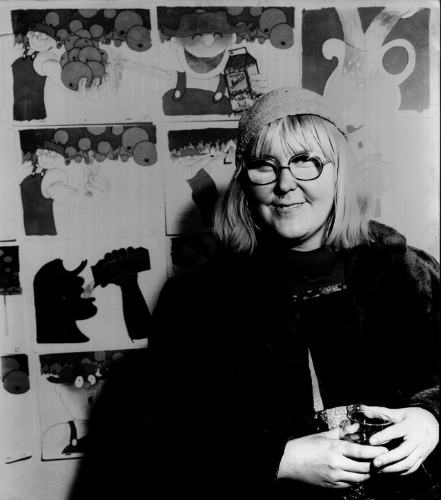 Black and white photograph of Marjut Rimminen, with a fair bob, glasses and a knitted cap, holding a glass in front of a wall of her Farmarin Juissi drawings, among them the farmer with a carton of Jussi juice.