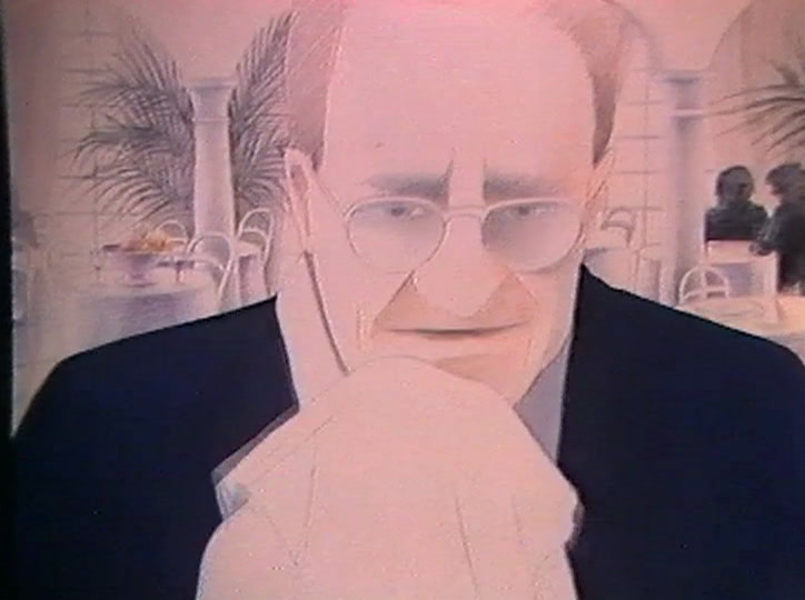 An older man in suit and glasses winces at a table in a glass-roofed restaurant