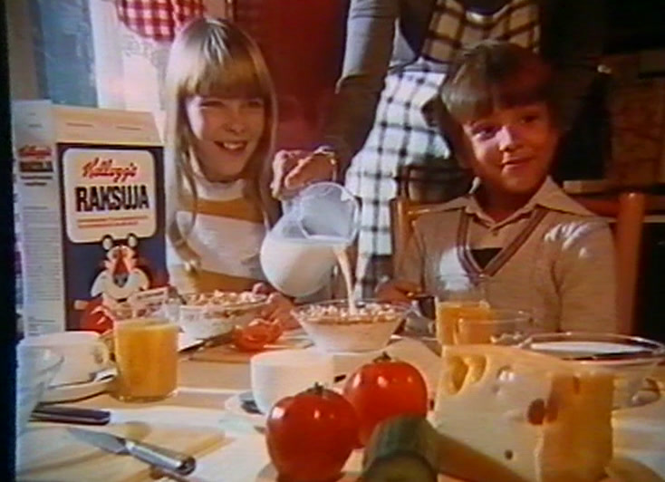 Two children at a laid table, milk being poured, a Raksuja pack behind them.