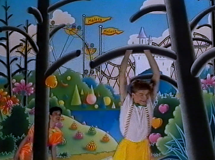 A woman in a yellow skirt stands on a painted sweet-strewn hillside behind dark tree trunks.