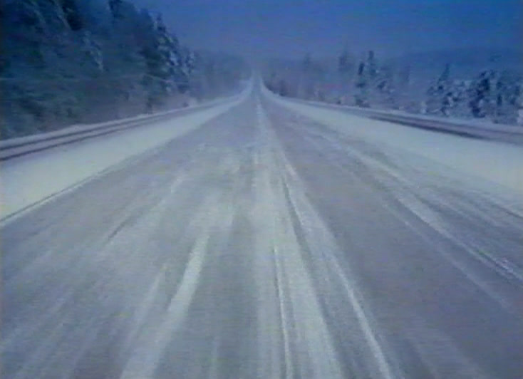 A snow-covered road runs straight through winter forest, filmed from a moving car.