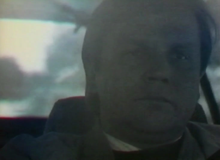 A man's face in the car, flat grey-green and nearly colourless.