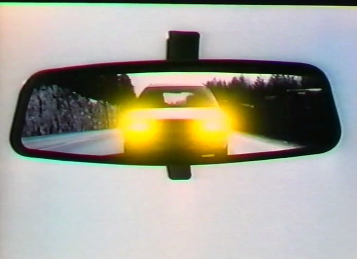 A rear-view mirror reflects a car's blazing yellow headlights on a snowy road.
