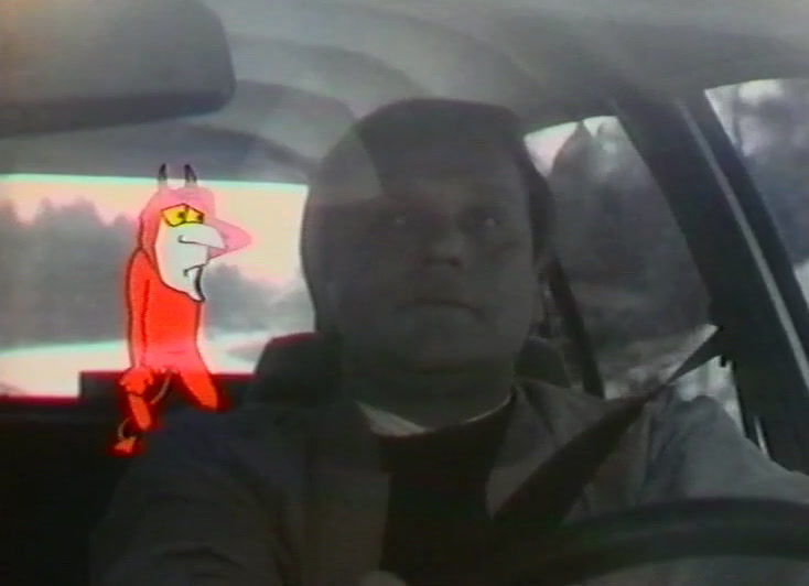 A small red devil stands on the dashboard beside the driver at the wheel.