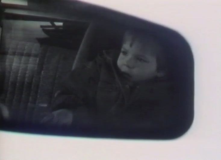 A wing mirror reflects a small child sitting in the back seat.