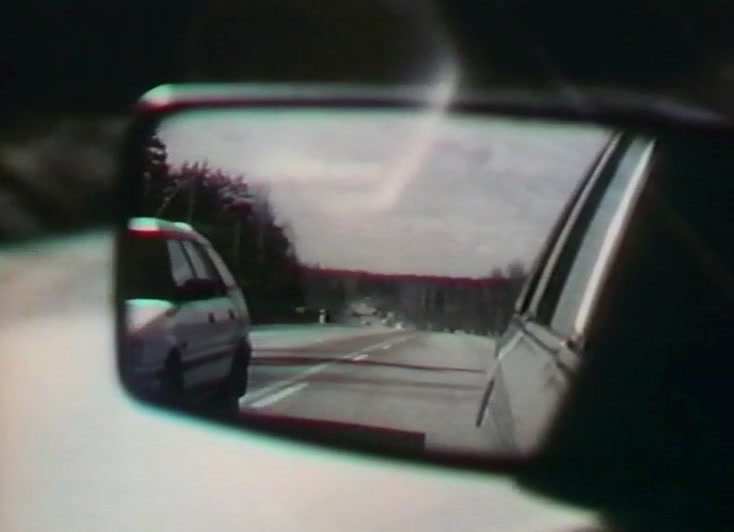 A wing mirror shows a car on the road behind, forest at the edge.