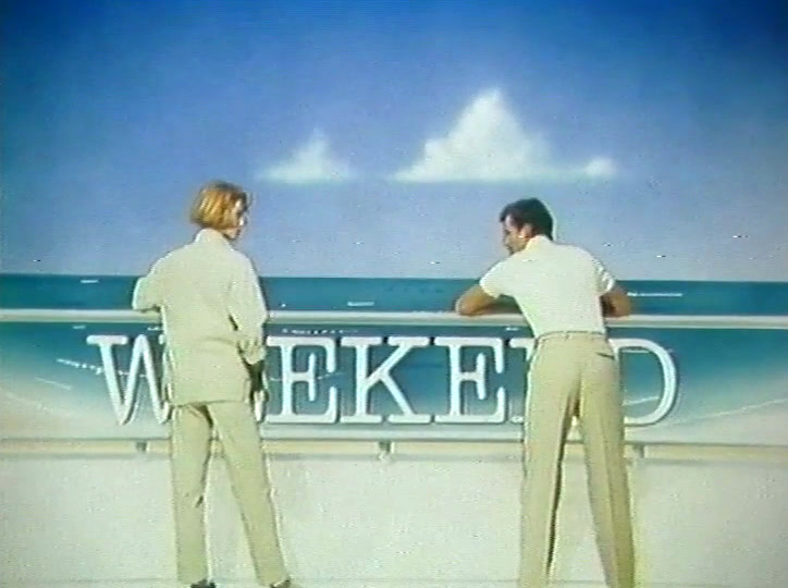 Seen from behind, two models in pale trousers face the painted sea horizon.