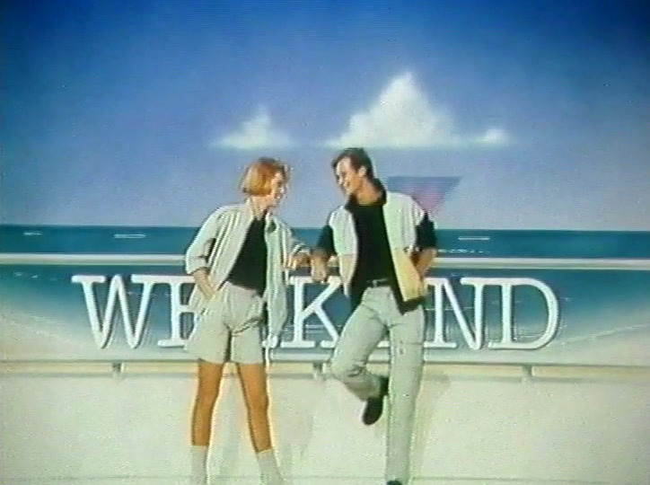 Two models in pale suits stand either side of the lettering, arms held out.